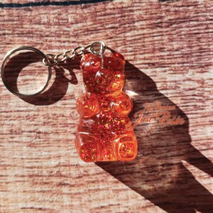 May include: A clear, orange gummy bear keychain with glitter embedded throughout. The keychain has a silver chain and ring. The background is a wooden surface. The text "Just Resin" is visible on the right side of the image.