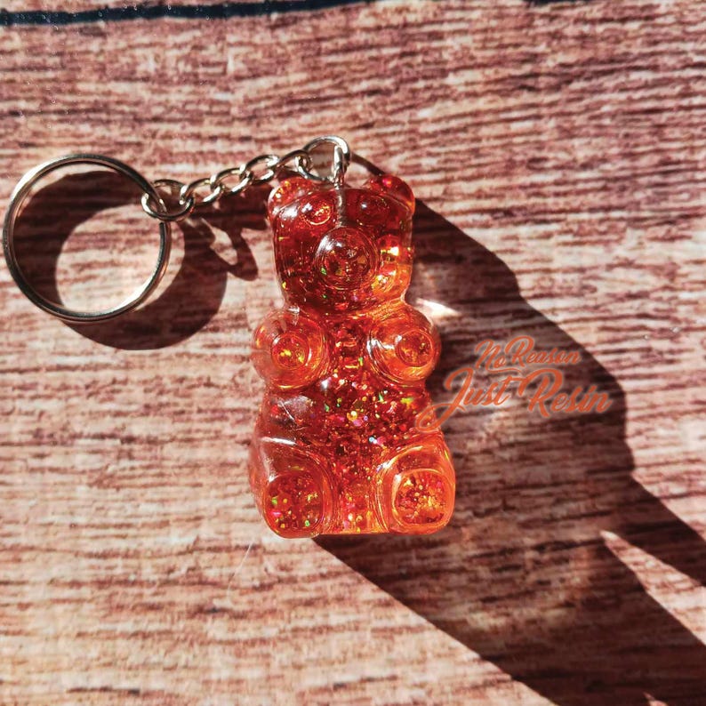 May include: A clear, orange gummy bear keychain with glitter embedded throughout. The keychain has a silver chain and ring. The background is a wooden surface. The text "Just Resin" is visible on the right side of the image.