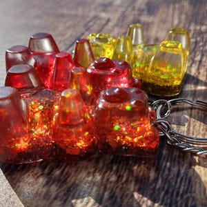 May include: A handmade resin keychain featuring a cluster of red and yellow gummy bear shapes. The bears are filled with glitter and attached to a silver keyring. The resin is translucent, allowing light to pass through.
