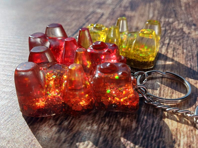 May include: A handmade resin keychain featuring a cluster of red and yellow gummy bear shapes. The bears are filled with glitter and attached to a silver keyring. The resin is translucent, allowing light to pass through.