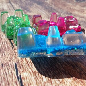 May include: A colorful resin art piece featuring a row of transparent, geometric shapes. The piece transitions from green to pink and blue, with glitter accents. The artwork is held by a hand, showcasing the vibrant colors and textures.