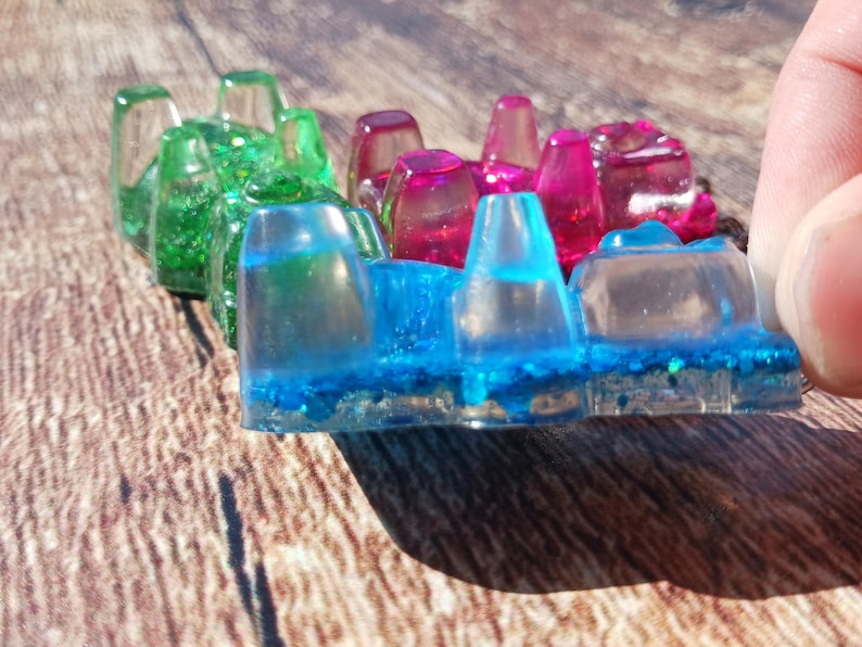 May include: A colorful resin art piece featuring a row of transparent, geometric shapes. The piece transitions from green to pink and blue, with glitter accents. The artwork is held by a hand, showcasing the vibrant colors and textures.