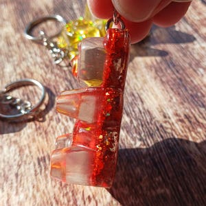 May include: A red glitter gummy bear keychain. The gummy bear is made of clear resin with red glitter and a metal keyring. The keychain is held up by a person's fingers. Other keychains are visible in the background.