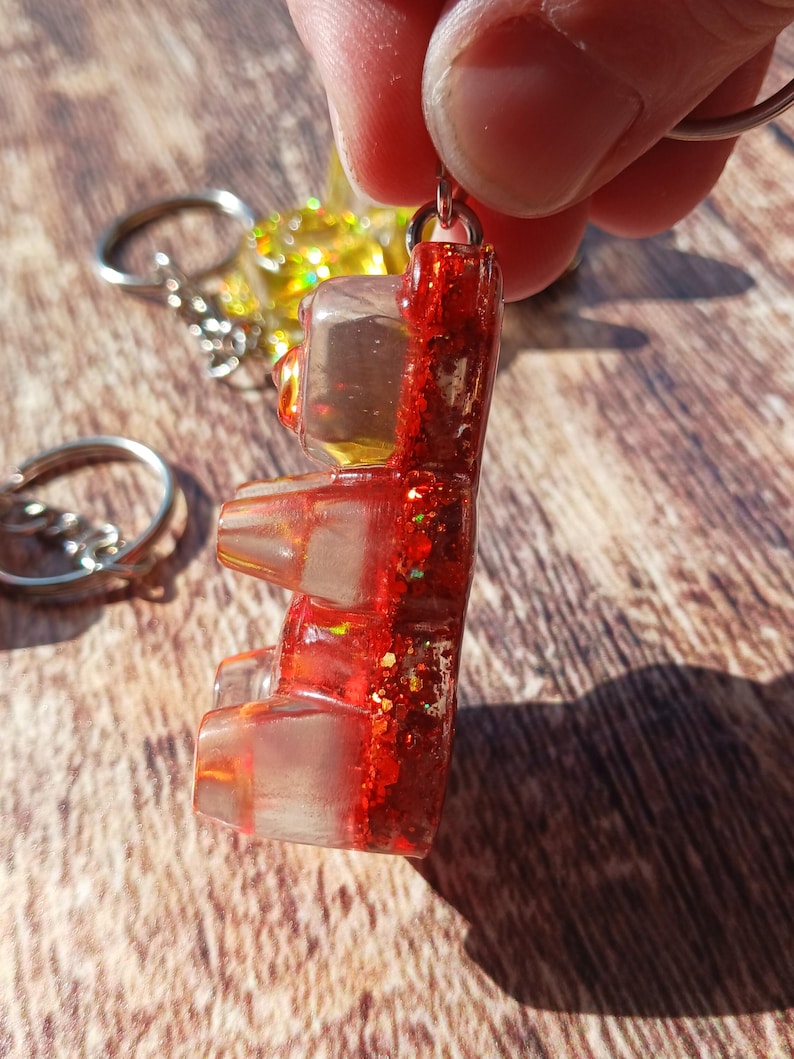 May include: A red glitter gummy bear keychain. The gummy bear is made of clear resin with red glitter and a metal keyring. The keychain is held up by a person's fingers. Other keychains are visible in the background.