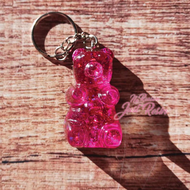 May include: A pink gummy bear keychain with a silver keyring and chain. The gummy bear is made of translucent resin with glitter embedded inside. The keychain is on a wooden surface.
