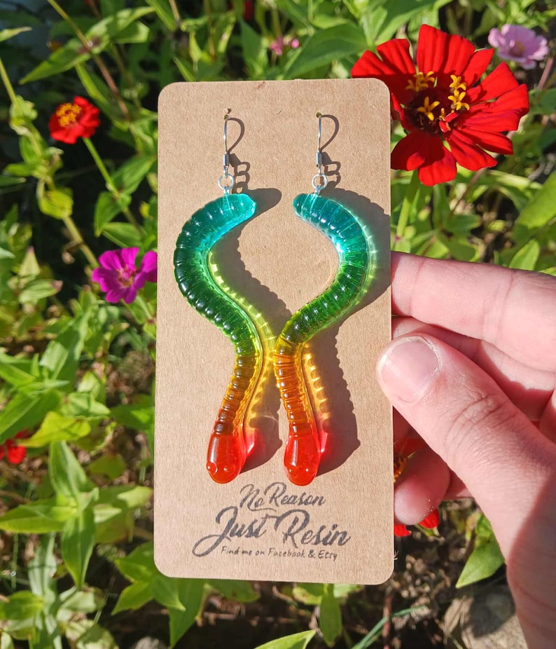 May include: A pair of colorful worm-shaped earrings. The earrings are made of resin and feature a gradient of colors, transitioning from red to yellow, green, and blue. The earrings are displayed on a brown card with the text "No Reason Just Resin".