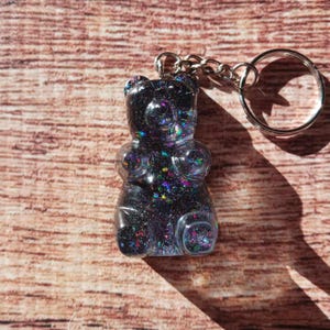 May include: A clear, glitter-filled gummy bear keychain. The bear is black with iridescent glitter, attached to a silver chain and keyring. The background is a wooden surface.