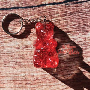 May include: A red, glitter-infused gummy bear keychain. The bear-shaped charm is attached to a silver keyring and chain. The keychain is made of a translucent red material with glitter embedded throughout. The background is a wooden surface.