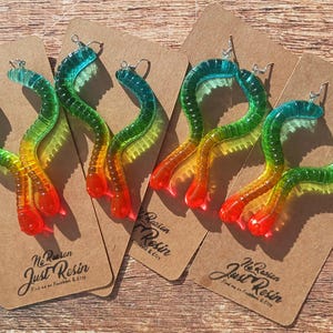 K&ouml;nnte beinhalten: Ohrringe in Wurmform aus Harz, in Farben von Blau bis Rot. Die Ohrringe werden auf braunen Pappkarten pr&auml;sentiert. Die Karten tragen den Text "Just Resin" und Social-Media-Informationen.