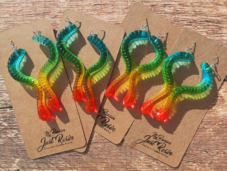 K&ouml;nnte beinhalten: Ohrringe in Wurmform aus Harz, in Farben von Blau bis Rot. Die Ohrringe werden auf braunen Pappkarten pr&auml;sentiert. Die Karten tragen den Text "Just Resin" und Social-Media-Informationen.