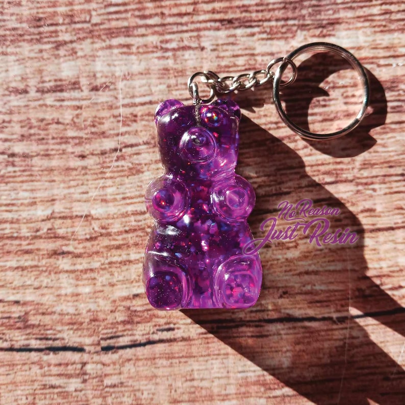 May include: A purple gummy bear keychain with glitter embedded in the resin. The keychain has a silver chain and a round key ring. The words "No Reason Just Resin" are in the bottom right corner.