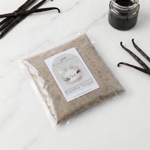 May include: A clear plastic bag filled with vanilla sugar, with a label that reads "Vanilla Sugar". The image also shows vanilla beans and a small jar of vanilla extract. The background is a white marble surface.