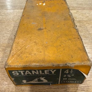 May include: A vintage Stanley hand plane in its original yellow box. The box features a green label with the Stanley logo and dimensions: 4 1/2, 2 3/8", and 60 mm. The box shows signs of wear and tear, indicating its age.