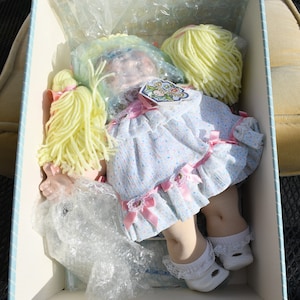 May include: A Cabbage Patch Kids doll in a white dress with pink bows and yellow yarn hair. The doll is in a box with bubble wrap.