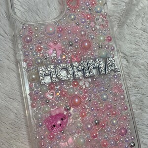 May include: Clear phone case with a pink, white and silver design. The case is covered in small beads and rhinestones. The word "MOMMA" is spelled out in silver rhinestones on the case.