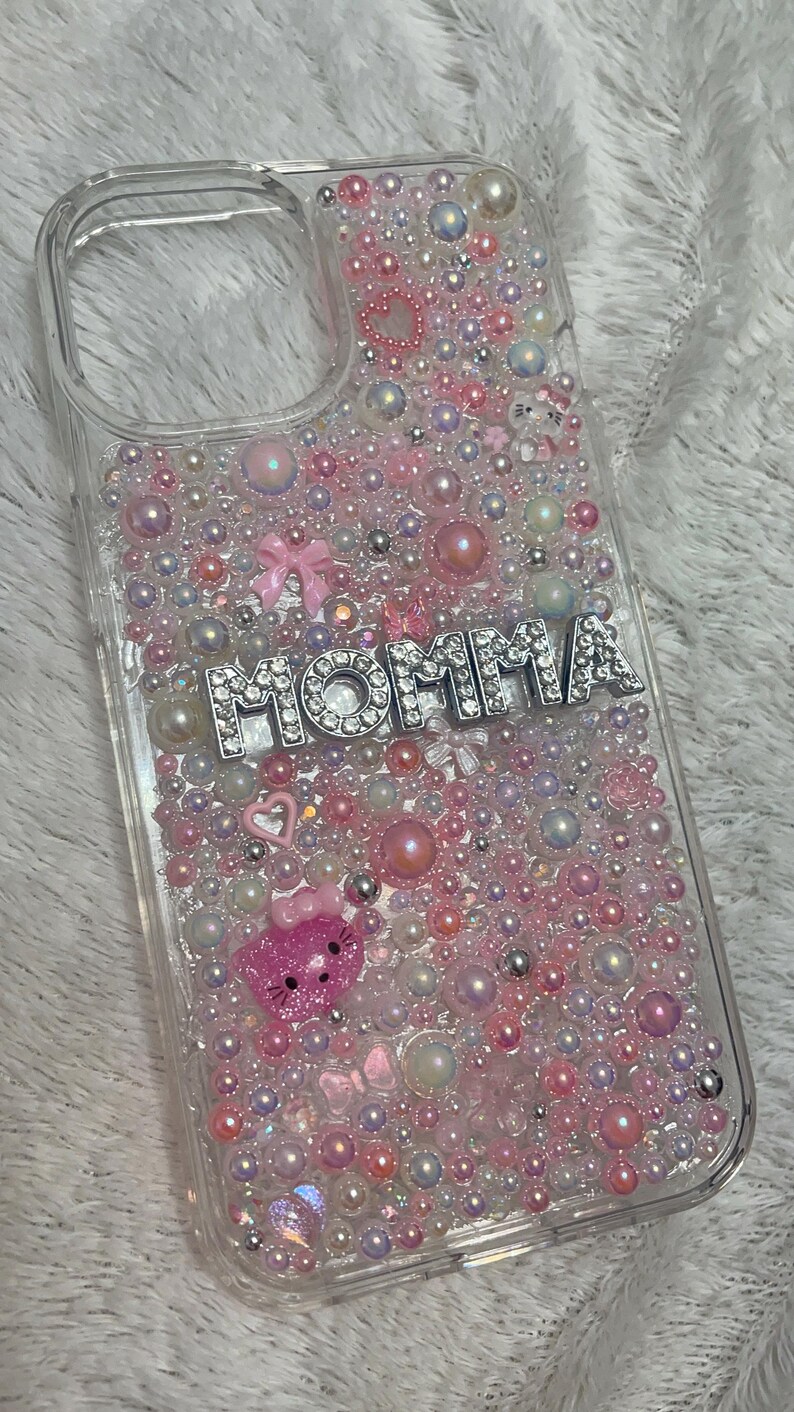 May include: Clear phone case with a pink, white and silver design. The case is covered in small beads and rhinestones. The word "MOMMA" is spelled out in silver rhinestones on the case.