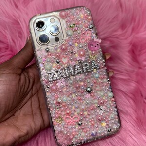 May include: A clear phone case with a pink, white and silver design. The case is covered in small beads and rhinestones. The name "ZAHARA" is spelled out in rhinestones on the case.