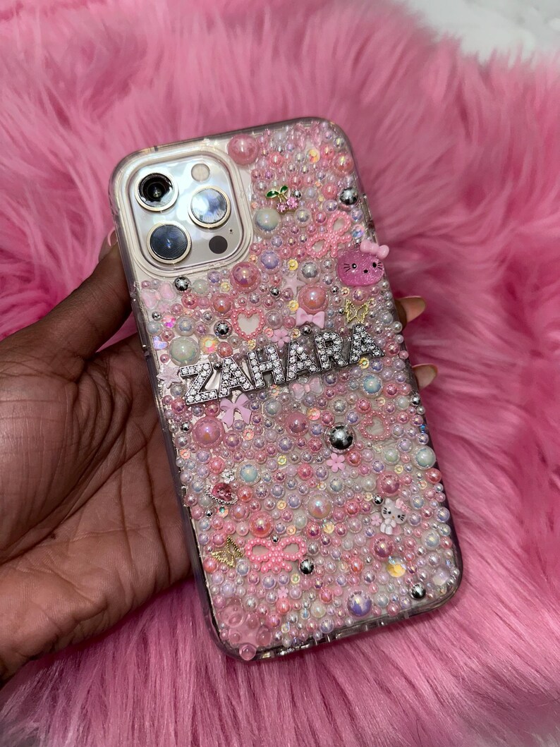 May include: A clear phone case with a pink, white and silver design. The case is covered in small beads and rhinestones. The name "ZAHARA" is spelled out in rhinestones on the case.