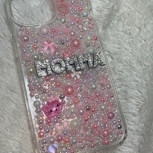 May include: A clear phone case with a pink, white and silver design. The case is covered in small pearls and rhinestones. The word "MOMMA" is spelled out in silver letters with rhinestones.