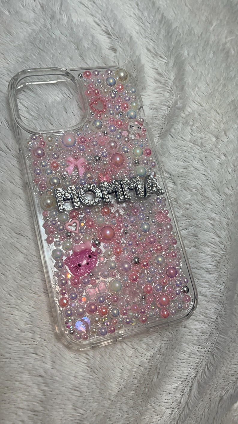 May include: A clear phone case with a pink, white and silver design. The case is covered in small pearls and rhinestones. The word "MOMMA" is spelled out in silver letters with rhinestones.