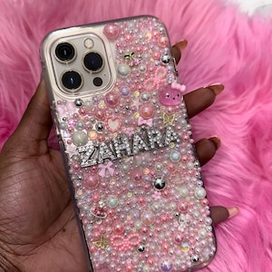May include: A clear phone case with a pink, white and silver design. The case is covered in small beads and rhinestones. The name "ZAHARA" is spelled out in silver rhinestones on the case.