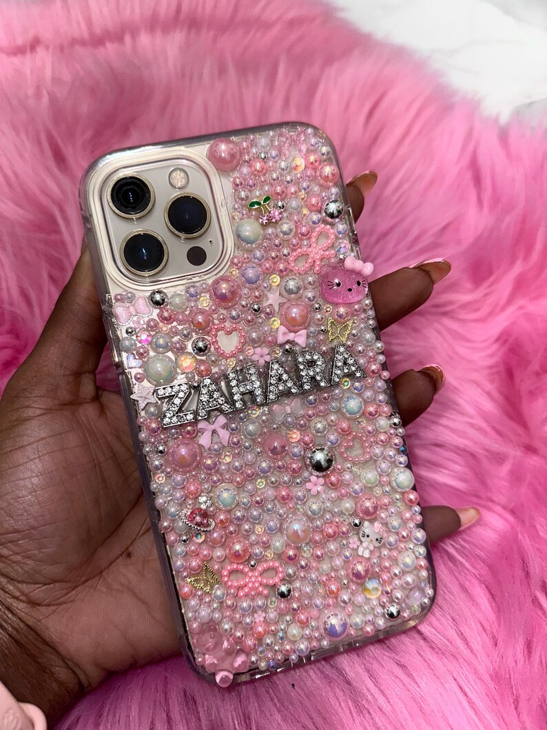 May include: A clear phone case with a pink, white and silver design. The case is covered in small beads and rhinestones. The name "ZAHARA" is spelled out in silver rhinestones on the case.