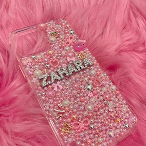 May include: A clear phone case with a pink, white and silver glitter design. The case has the name "Zahara" spelled out in silver rhinestones.