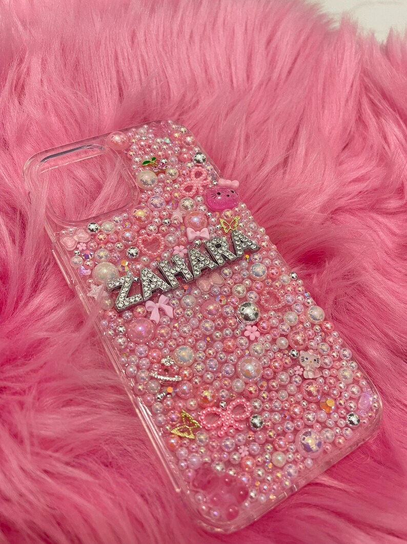 May include: A clear phone case with a pink, white and silver glitter design. The case has the name "Zahara" spelled out in silver rhinestones.