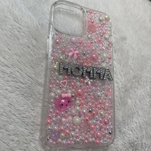 May include: A clear phone case with a pink, white and silver design. The case is covered in small beads and has the word "MOMMA" written in silver letters. There is a pink Hello Kitty character on the case.