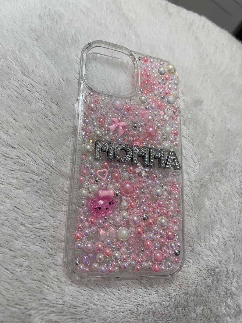 May include: A clear phone case with a pink, white and silver design. The case is covered in small beads and has the word "MOMMA" written in silver letters. There is a pink Hello Kitty character on the case.
