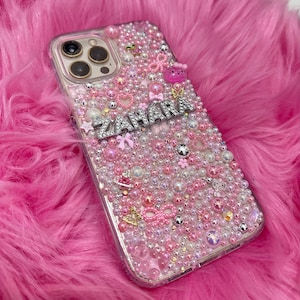 May include: A clear phone case with a pink, white and silver design. The case is covered in small beads and rhinestones. The name "ZAHARA" is spelled out in silver rhinestones on the case.