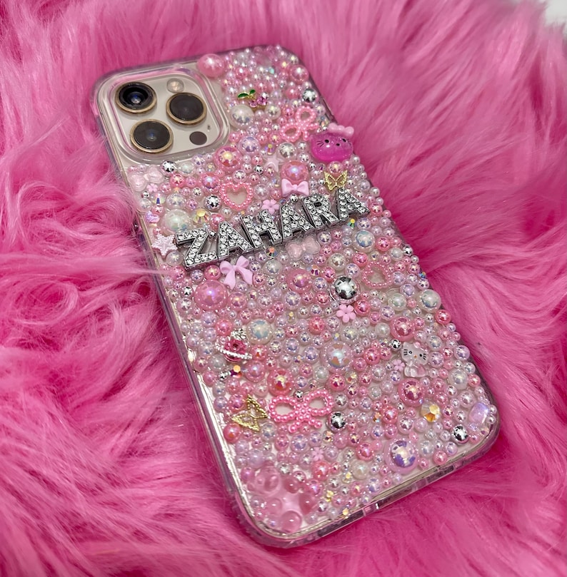 May include: A clear phone case with a pink, white and silver design. The case is covered in small beads and rhinestones. The name "ZAHARA" is spelled out in silver rhinestones on the case.