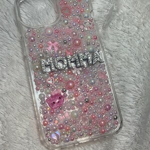 May include: A clear phone case with a pink, white and iridescent glitter design. The case has a silver charm that says "MOMMA" with rhinestones.