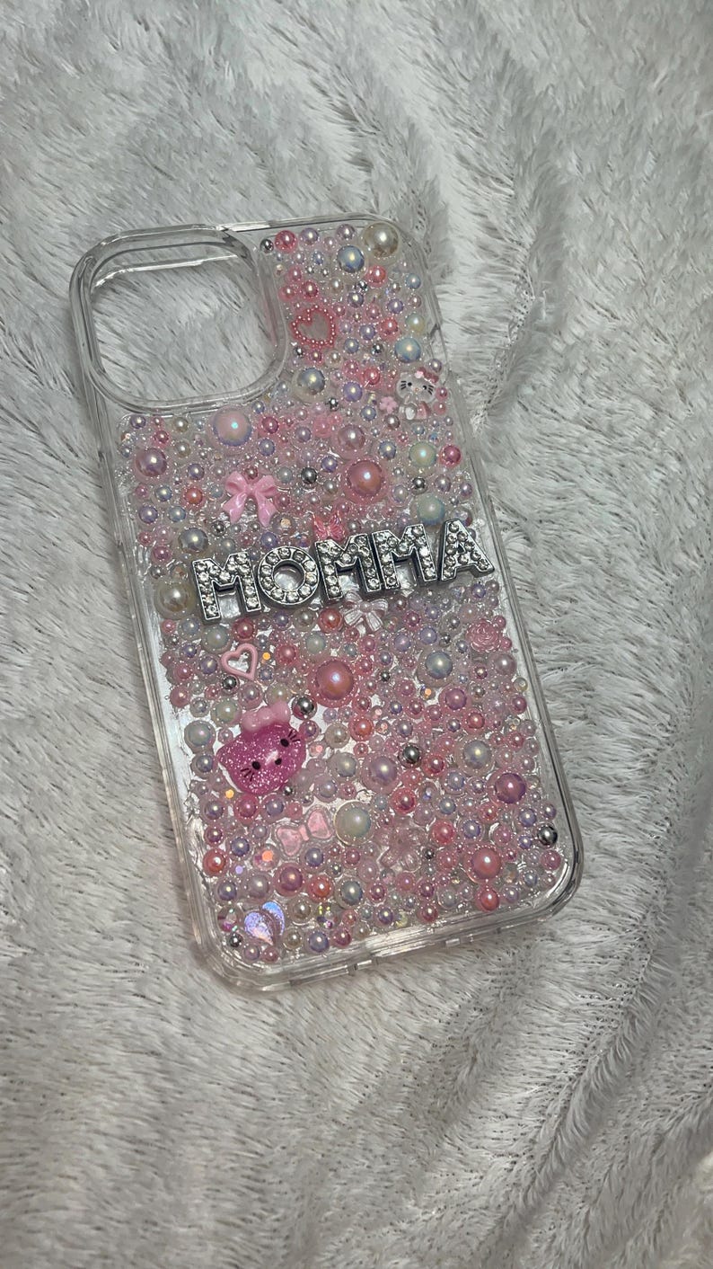 May include: A clear phone case with a pink, white and iridescent glitter design. The case has a silver charm that says "MOMMA" with rhinestones.