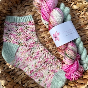 May include: A knitted sock with a floral design in pink, white, and green, next to two skeins of hand-dyed yarn in matching colours. The yarn is labelled "Lorna Campbell Designs". The items are presented in a woven basket.