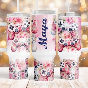 Puede incluir: Un vaso de acero inoxidable blanco con un diseño floral rosa y blanco y un mando de videojuego. El vaso tiene un asa y una tapa con una pajita. El nombre "Maya" está escrito en el vaso.