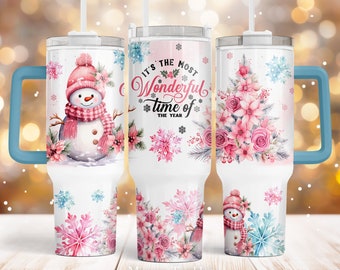 May include: A white stainless steel tumbler with a blue handle. The tumbler features a pink and white winter scene with a snowman, snowflakes, and flowers. The text "It's the most Wonderful time of the year" is printed on the tumbler.