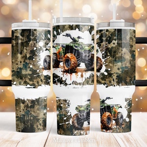 Puede incluir: Un vaso de acero inoxidable con asa negra y tapa. El vaso presenta un patrón de camuflaje verde y marrón con salpicaduras de pintura blanca. Un ATV verde está en el centro del diseño, conduciendo por el barro. El texto "Mamaya Tumblers" está en la parte inferior de la imagen.