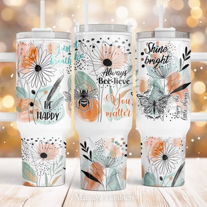 Könnte beinhalten: Drei Edelstahlbecher mit Blumendesigns und inspirierenden Zitaten. Die Becher haben einen weißen Hintergrund mit schwarzen Umrissen von Blumen, Bienen und Schmetterlingen. Die Zitate lauten "Atme einfach", "Glaub immer an dich", und "Strahle, genieße die kleinen Dinge".