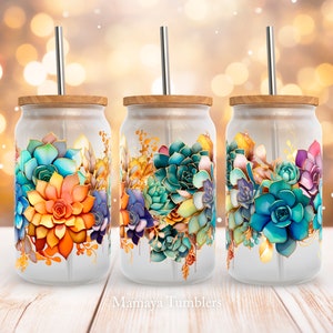 May include: Three clear glass tumblers with wooden lids and metal straws. Each tumbler features a different arrangement of colorful succulent plants with gold accents. The tumblers are arranged on a wooden surface. The text "Mamaya Tumblers" is visible below the tumblers.