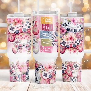 May include: A white stainless steel tumbler with a pink and white floral design and a pink and white video game controller. The tumbler has a handle and a straw. The text "Gamer Girl" is on the tumbler.