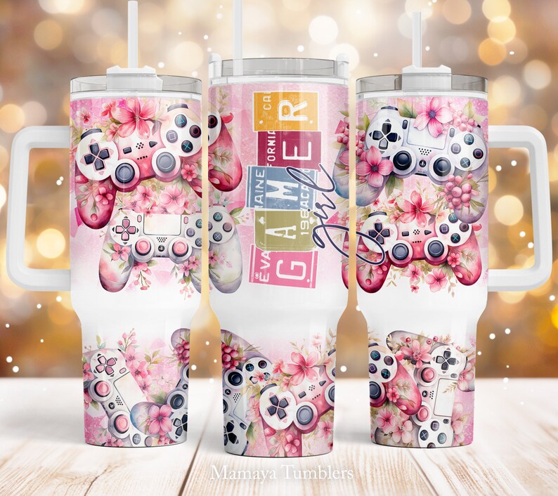May include: A white stainless steel tumbler with a pink and white floral design and a pink and white video game controller. The tumbler has a handle and a straw. The text "Gamer Girl" is on the tumbler.