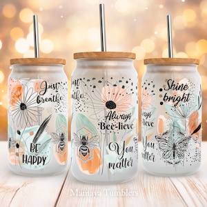 May include: Three glass tumblers with wooden lids and stainless steel straws. Each tumbler features a different floral design and inspirational quote. The quotes are "Just breathe", "Always Bee-lieve", and "Shine bright".