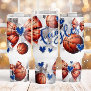 May include: White tumbler with a basketball and bow design. The design includes red bows, basketballs, blue hearts, and the word "Eagles" in blue script. The tumbler has a white handle and a straw.