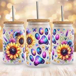 Op de afbeelding: Drie witte bekers met een galaxy-pootafdruk ontwerp en een bloemenontwerp met zonnebloemen. De bekers hebben een houten deksel en een rietje.