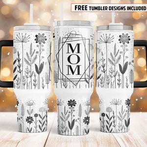 May include: White stainless steel tumbler with a black handle and lid. The tumbler features a black and white design of flowers and leaves with the word "MOM" in a geometric frame.
