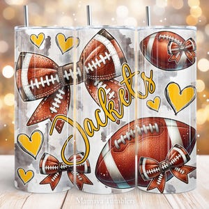 May include: White tumbler with a watercolor background featuring a brown football with white laces, a brown bow with white stitching, and the word "Jackets" in yellow script. There are also yellow hearts and smaller brown bows with white stitching.