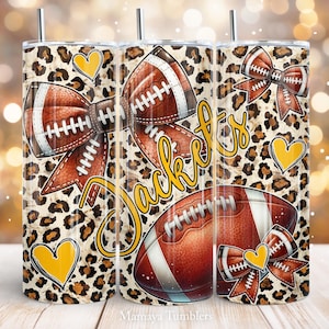 May include: A set of three stainless steel tumblers with a leopard print background. The tumblers feature a football, a bow, and the word "Jackets" in yellow lettering. The bow is made of footballs and has a yellow heart in the center.