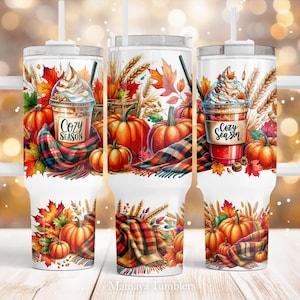 Puede incluir: Un vaso blanco con un diseño de otoño. El vaso presenta una bufanda de cuadros, hojas de otoño y calabazas. El texto "Cozy Season" está impreso en el vaso.