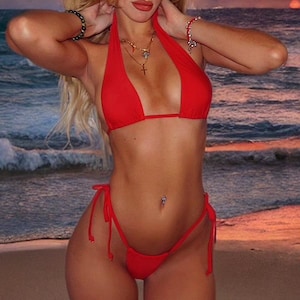 Op de afbeelding: Een rode halterbikini set. De top heeft een strik in de nek en op de rug, en de broekjes hebben strikzijden. Het model staat op een zandstrand met de oceaan en een zonsondergang op de achtergrond.